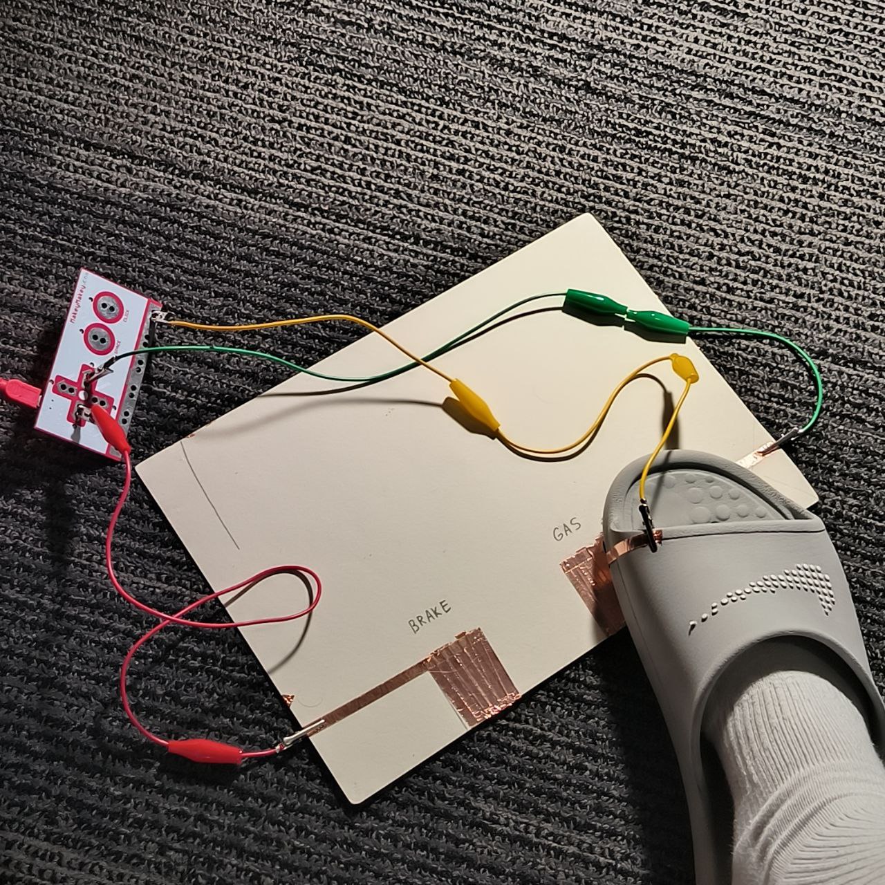 Finally, the slipper makes contact with one of the copper zones, likely sending an input signal to the Makey Makey. The setup simulates a foot-controlled interface, like pressing gas or brake in a game or sound-based sketch. 